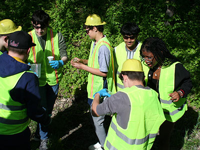 A group of students dressed in safety vests and other protective gear gather outside with cups in hand preparing to test ground water