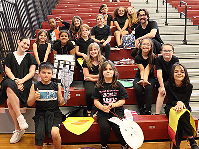 Drama Club students dressed mostly in black sit on the bleachers with club advisers and smile at the camera