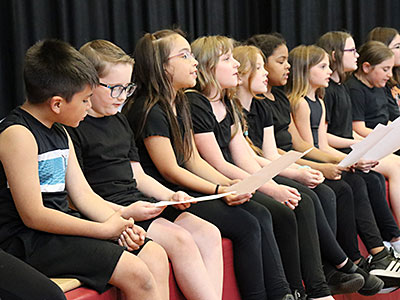 A group of students sit at the edge of the stage holding music sheets and singing