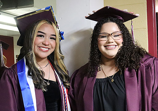 Two graduates dressed in caps and gowns smile as they stand together