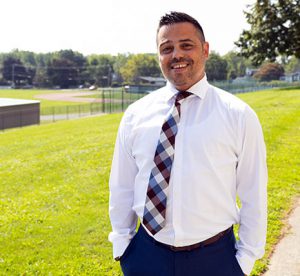 Watervliet Junior Senior High School assistant principal dressed in white shirt and tie stands outside the school building with baseball field and tennis courts in the background.