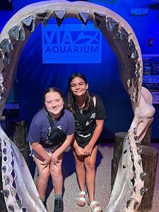 Two students pose behind a giant pair of shark jaws at the Via Aquarium during a summer travel field trip