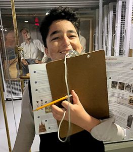 A student holds a clipboard and smiles in front of an exhibit during a summer travel field trip to FDR's estate in Hyde Park