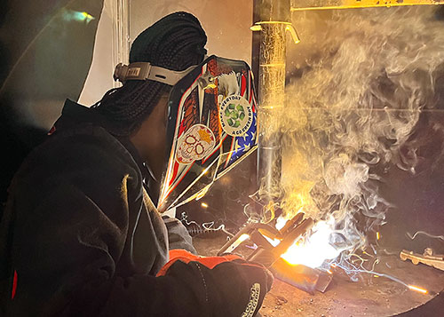 Student dressed in helmet and other protective gear holds welding torch that is sparking