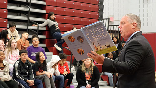 Assembly member McDonald holds a book in his left had and reads to students who are seated in the bleachers in the gymnasium