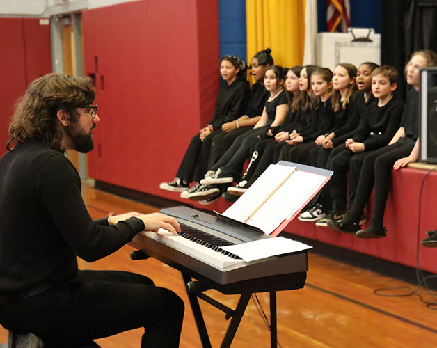 Drama Club adviser plays keyboard as cast members sit on edge of stage joined together in song