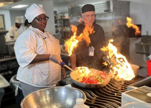 Culinary student and chef teacher stand at a stove watching as flames leap from a large frying pan 